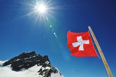 Low angle view of swiss flag by snow covered mountain against bright sun
