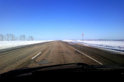 Empty road by snow covered field against clear blue sky seen through car windshield