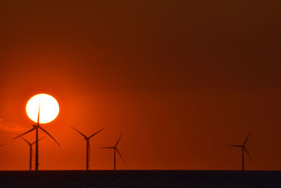 Silhouette wind turbines in sea against orange sky