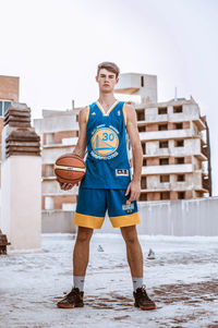 Portrait of young man holding basketball while standing against building