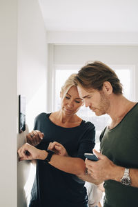 Mature couple adjusting smart watch with digital tablet mounted on wall at home