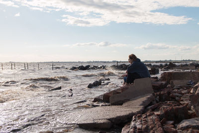 Man sitting on rock by sea against sky