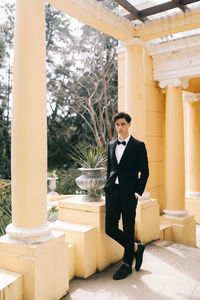 A beautiful young man, the groom in an elegant wedding suit, stands posing in the city's old park