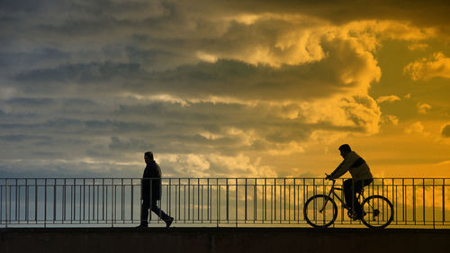 Man riding bicycle on bridge against cloudy sky during sunset