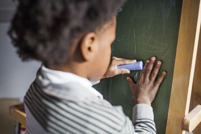 Rear view of boy writing with chalk in blackboard at home