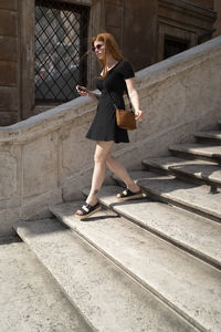 Red ginger woman in black dress descending stairs holding a smartphone