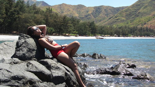 Sensuous woman in bikini leaning on rock formation by lake against mountains