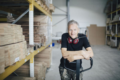 Portrait of smiling worker leaning on pallet jack by stack at industry