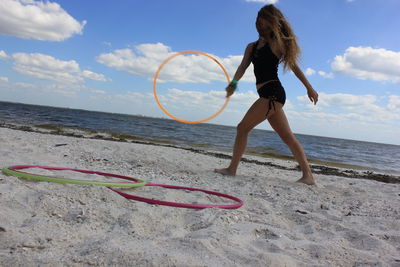 Full length of young woman playing with plastic hoop at beach against cloudy sky