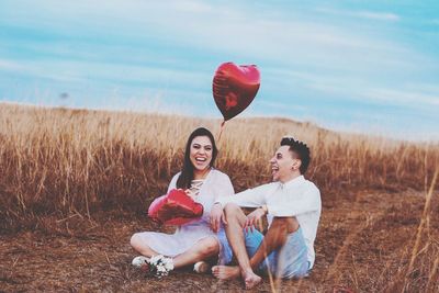Happy couple with heart shape balloon sitting on field against sky