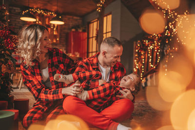 Smile active parents with small son in red checkered sleepwears waiting santa indoor.