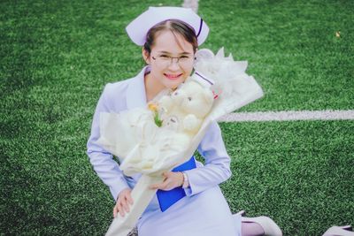 Portrait of smiling young woman in nurse uniform holding teddy bear while sitting on grassy field