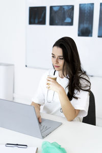 Young female doctor having coffee while using laptop in clinic