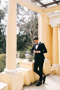 A beautiful young man, the groom in an elegant wedding suit, stands posing in the city's old park