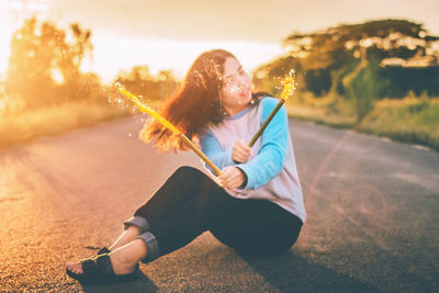 Woman holding illuminated firework while sitting on road during sunset