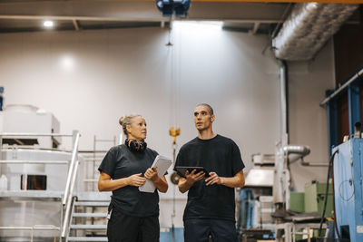 Front view of male worker holding tablet pc while discussing with female coworker in manufacturing warehouse