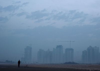 Man standing on street by buildings against cloudy sky in foggy weather