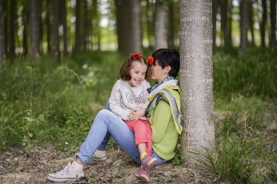 Smiling cute daughter sitting on lap of mother at forest