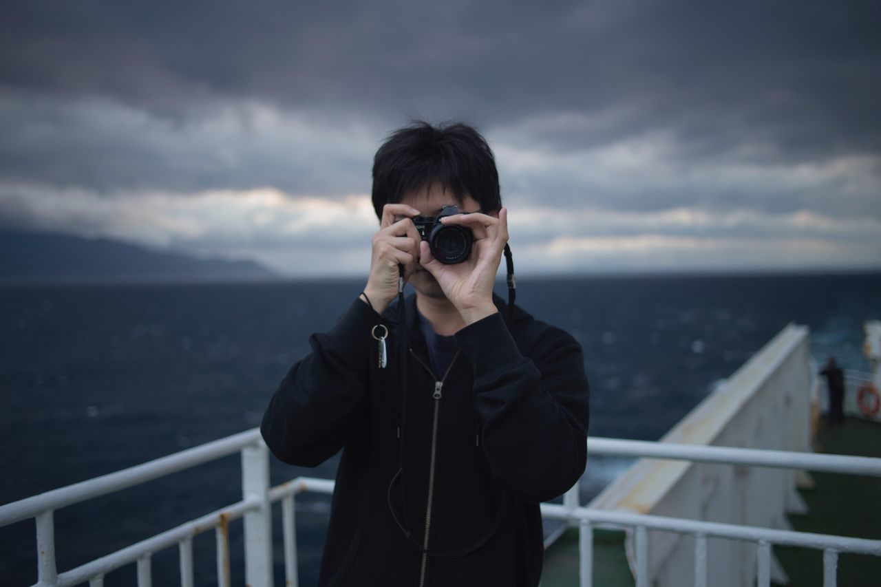 sea, cloud - sky, waist up, railing, sky, photography themes, real people, standing, one person, leisure activity, photographing, outdoors, camera - photographic equipment, front view, water, nature, horizon over water, holding, lifestyles, day, technology, photographer, young adult, young women, people