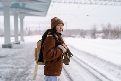 Young pensive asian woman with backpack standing at snow-covered railway platform in winter