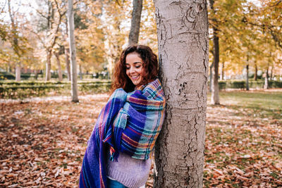 Smiling woman wrapped in blanket standing by tree trunk during autumn