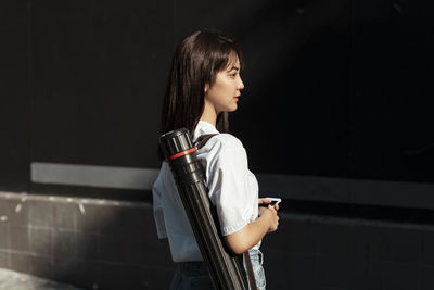 Side view of young woman using phone while standing against wall