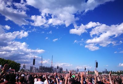 Crowd with arms raised standing in city during music concert against cloudy sky