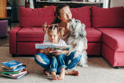 Mom reading book with baby girl toddler at home. funny home lifestyle family with licking pet dog. 