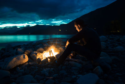 Man sitting by bonfire against cloudy sky at dusk