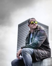 Low angle view of mid adult man wearing sunglasses while sitting against sky in city