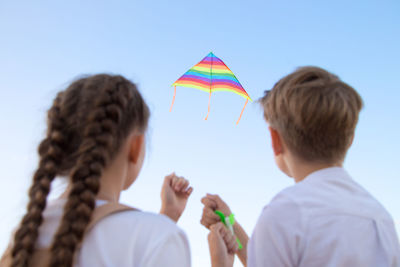 A girl and a boy launch a bright kite into the sky view from the back.