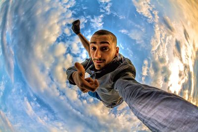 Fish-eye lens portrait of young man showing thumbs up amidst clouds