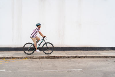 Full body side view of active male in protective helmet riding bicycle on sidewalk along road on street of city