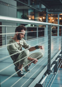 Side view of thoughtful handsome mid adult man looking away while crouching by railing in building