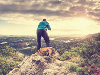 Tall photographer with baseball cap and tripod with camera in hands stand on rock and watch down