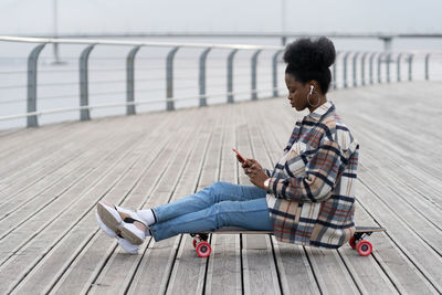 Young african girl sit on longboard with smartphone in hand text outdoor in park using 5g connection