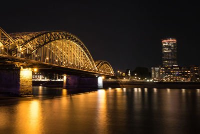 Illuminated bridge over river by buildings against sky at night