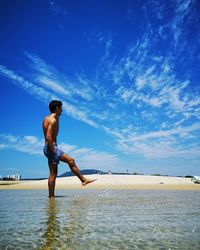 Full length of shirtless man talking on mobile phone while splashing water at beach against sky