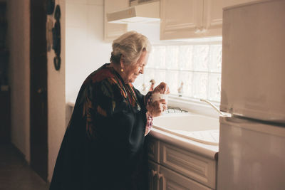 Side view of elderly female in warm shawl with mug standing near white sink in light white kitchen