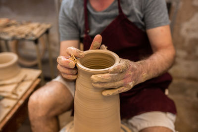 Crop unrecognizable sculptor with equipment giving shape while sculpting with brown clay on throwing wheel