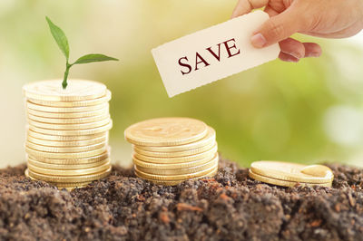 Cropped hand holding paper with save text over coins on dirt