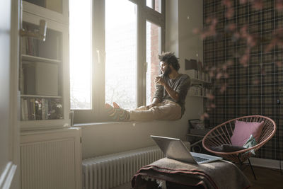 Man sitting on window sill in living room looking outside holding a cup