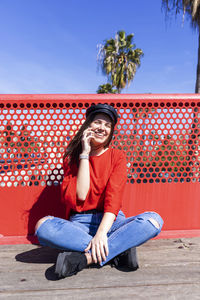 Teenage girl using mobile phone while wearing red top in city against railing during sunny day