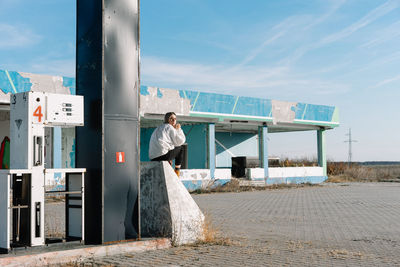 Young woman sitting at fuel pump against sky