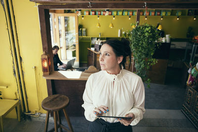 High angle view of female owner holding digital tablet while looking away in cafe