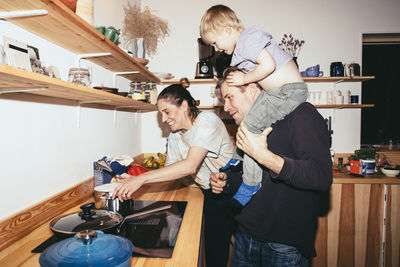 Father carrying son on shoulder by woman cooking food in kitchen at home