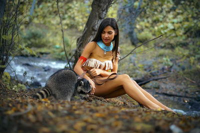 Smiling young woman in traditional clothing sitting by raccoon in forest