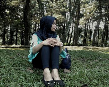 Thoughtful young woman wearing hijab while sitting on grassy field against trees