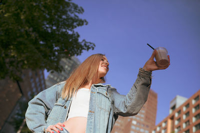 Low angle view of young woman holding drink glass against clear sky
