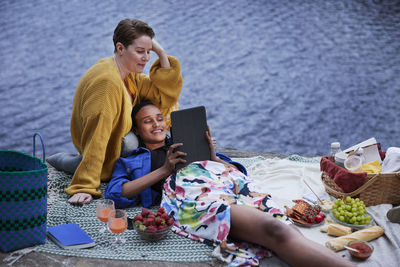 Female couple having picnic by river and using tablet